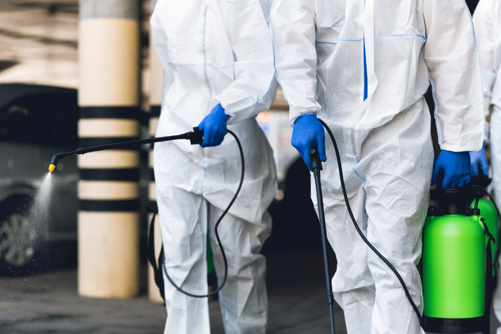 Two Professional Cleaners in Full Protective Suits and Gloves Performing Decontamination Using Spray Equipment in a Controlled Environment